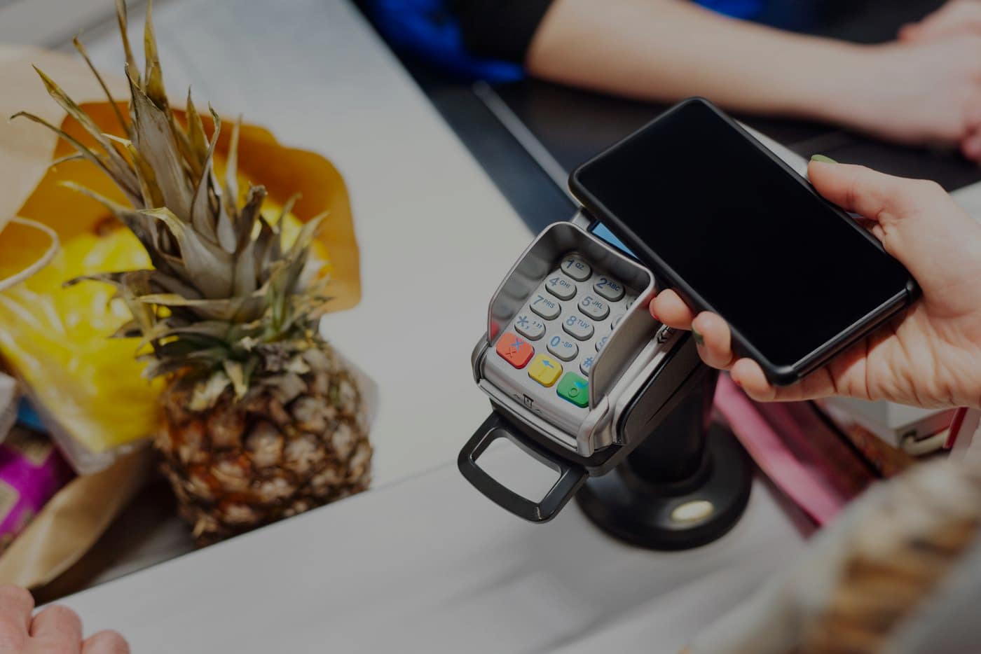 A phone held against a card terminal at a supermarket checkout, shopping on the belt beside it.